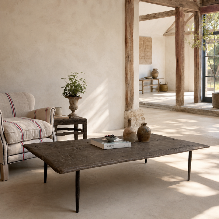 A rustic dark wood coffee table with black legs in a bright, sunlit farmhouse-style living room. A striped armchair, a small side table with a potted plant, and rustic ceramic vases are arranged around the table.