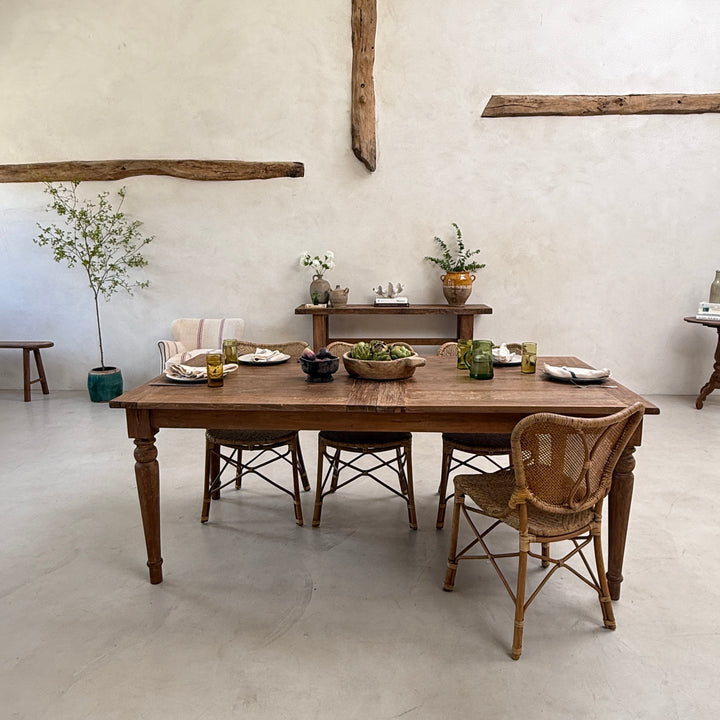 A rustic wooden dining table set with four woven and upholstered chairs in a bright, minimalist room with plaster walls and exposed wooden beams.