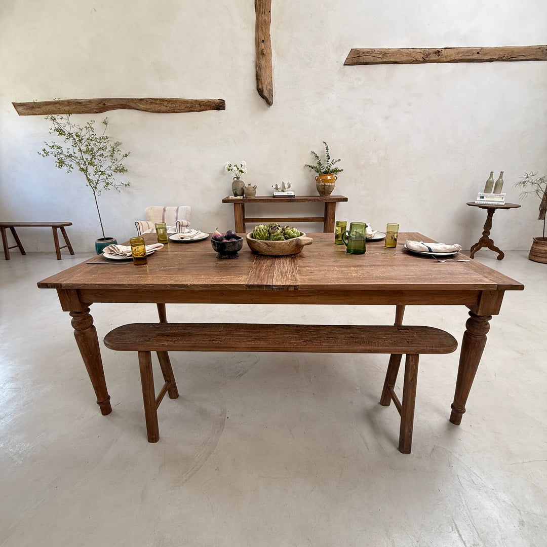 A rustic wooden dining table with a matching bench, set in a bright, minimalist room with white walls and exposed wood beams. The table is styled with plates, green glassware, and a bowl of grapes.