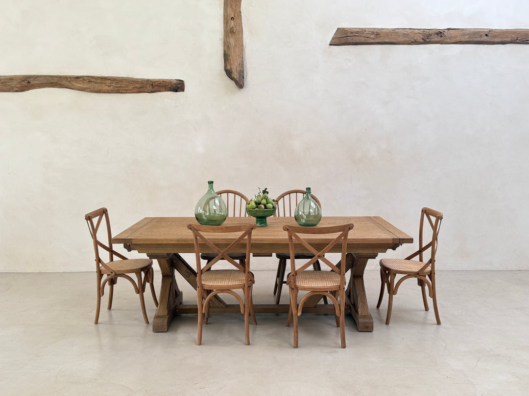 Reclaimed oak farmhouse dining table styled with six wooden chairs and simple green glass vases against a pale plaster wall.