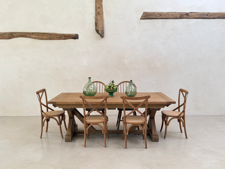 Reclaimed oak farmhouse dining table styled with six wooden chairs and simple green glass vases against a pale plaster wall.