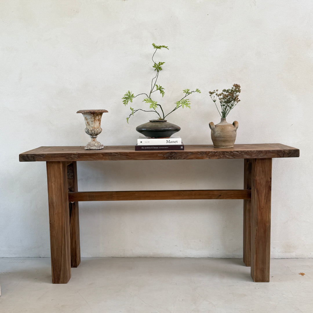 The Reclaimed Teak Console Table styled with handcrafted ceramics, antique stoneware and carefully chosen decorative objects against a softly textured plaster wall. Crafted from beautifully salvaged teak with a richly characterful grain and substantial architectural legs, this handcrafted console table brings understated elegance to entrance halls, living rooms and contemporary country interiors.
