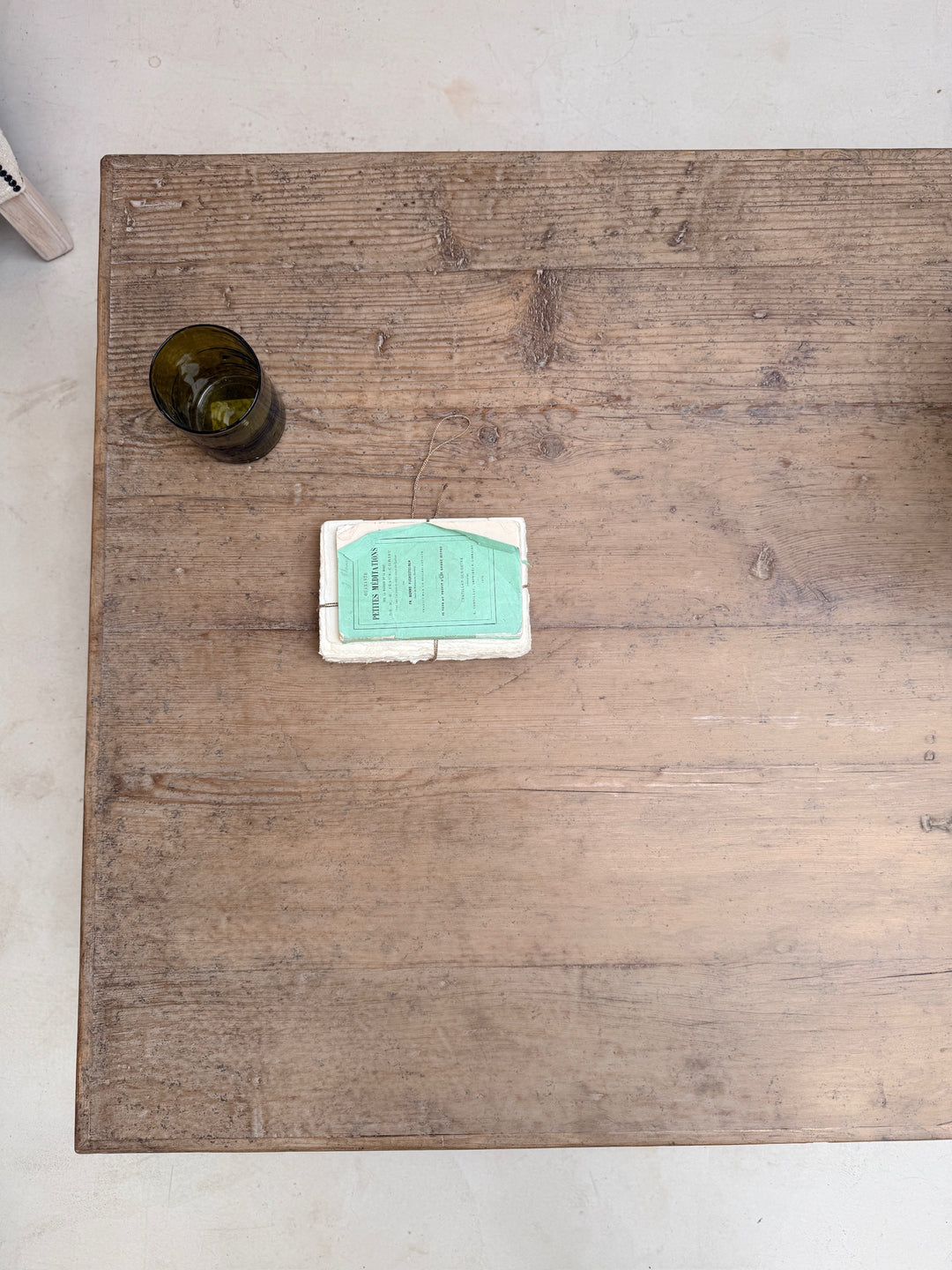 A rustic rectangular wooden table with a weathered brown finish and visible grain, photographed from above on a light floor. A small dark glass and a white box with a mint-green card are placed on the tabletop.