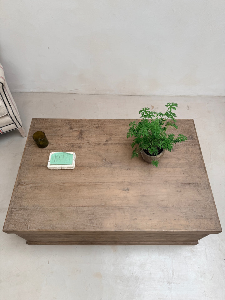 A rectangular wooden coffee table with a rustic brown finish, styled with a small green potted plant and a white container on top, set against a plain off-white wall and floor.