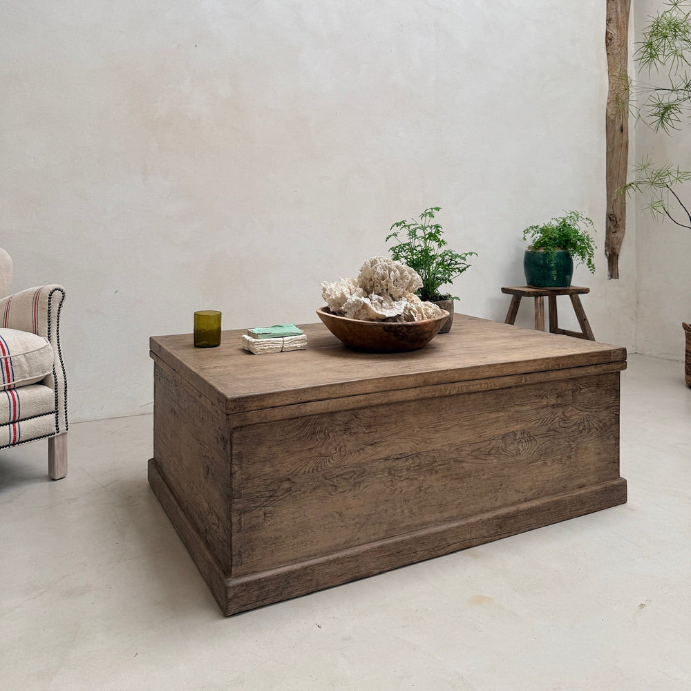 A rustic wooden chest coffee table in a minimalist room with a textured white wall, a wooden beam, and small plants on a side stool.