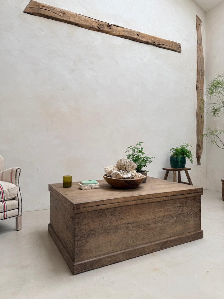 A rustic wooden chest coffee table in a minimalist room with a textured white wall, a wooden beam, and small plants on a side stool.