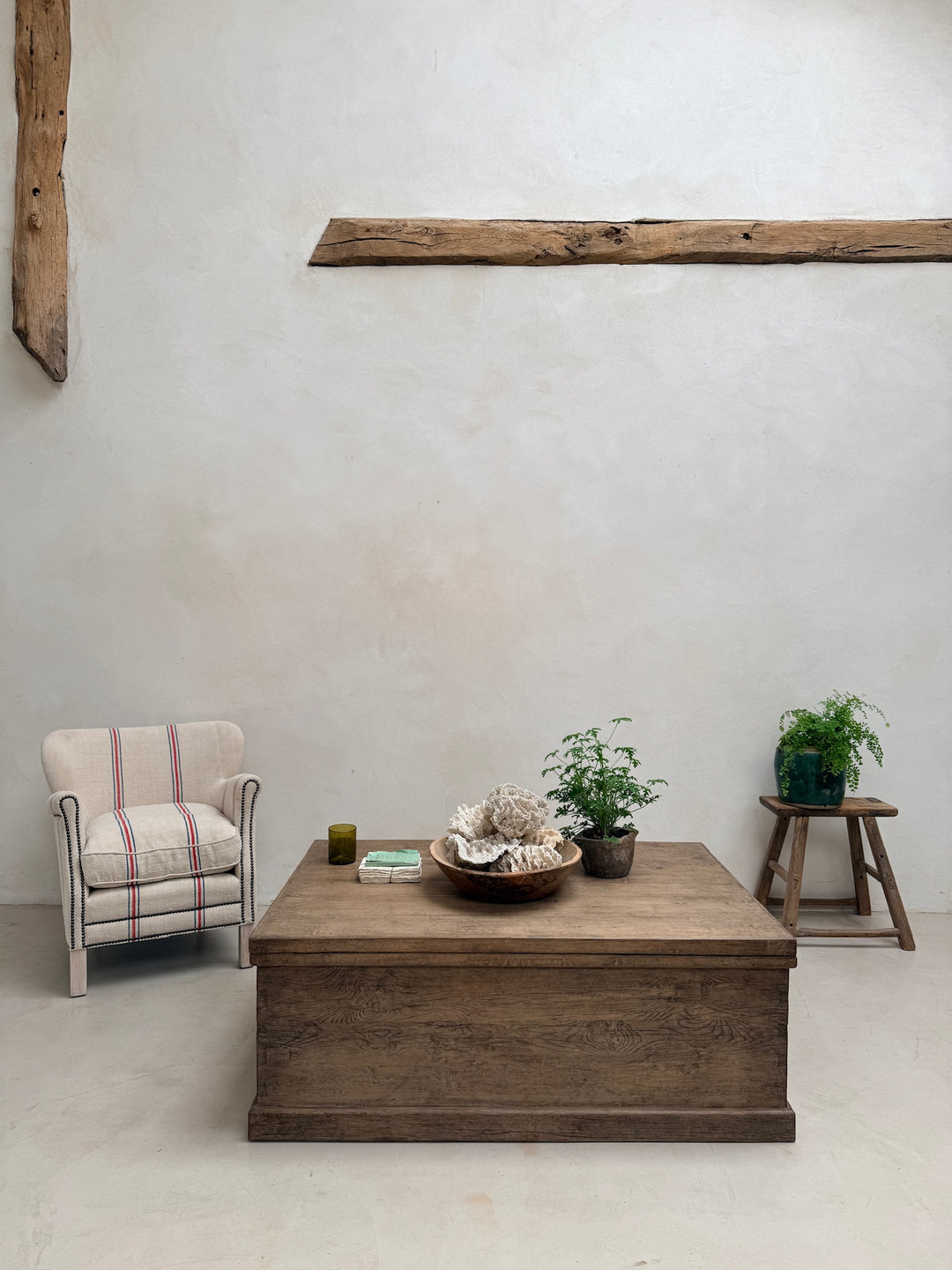 A rustic wooden coffee table in the center of a minimalist room, styled with a striped armchair, a small wooden stool with a potted plant, and decorative objects on top, set against a textured off-white wall with a horizontal wooden beam.