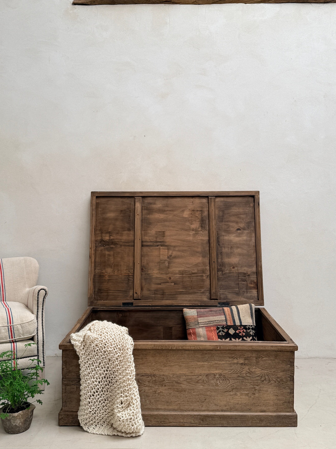A rustic dark brown wooden storage chest with its lid open, styled with a cream knit throw blanket and patterned cushions, set against a textured off-white wall.