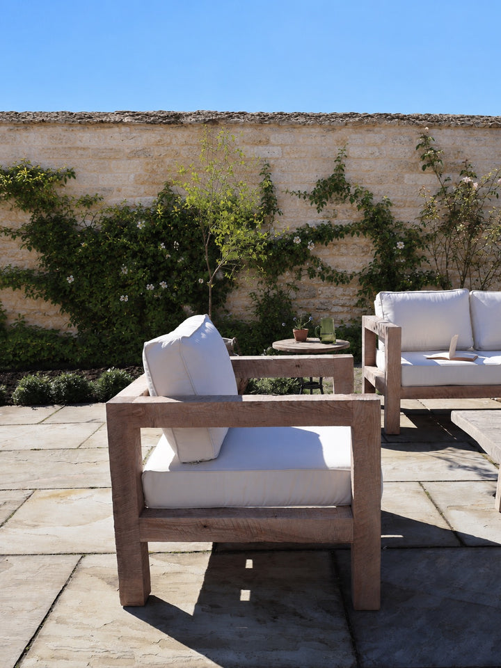 Wooden outdoor furniture with white cushions on a patio with a stone wall and greenery in the background.