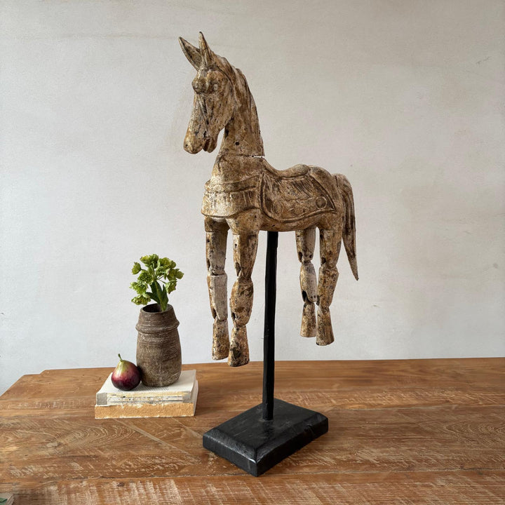 Wooden horse figurine on a stand with a plant and book on a wooden surface.