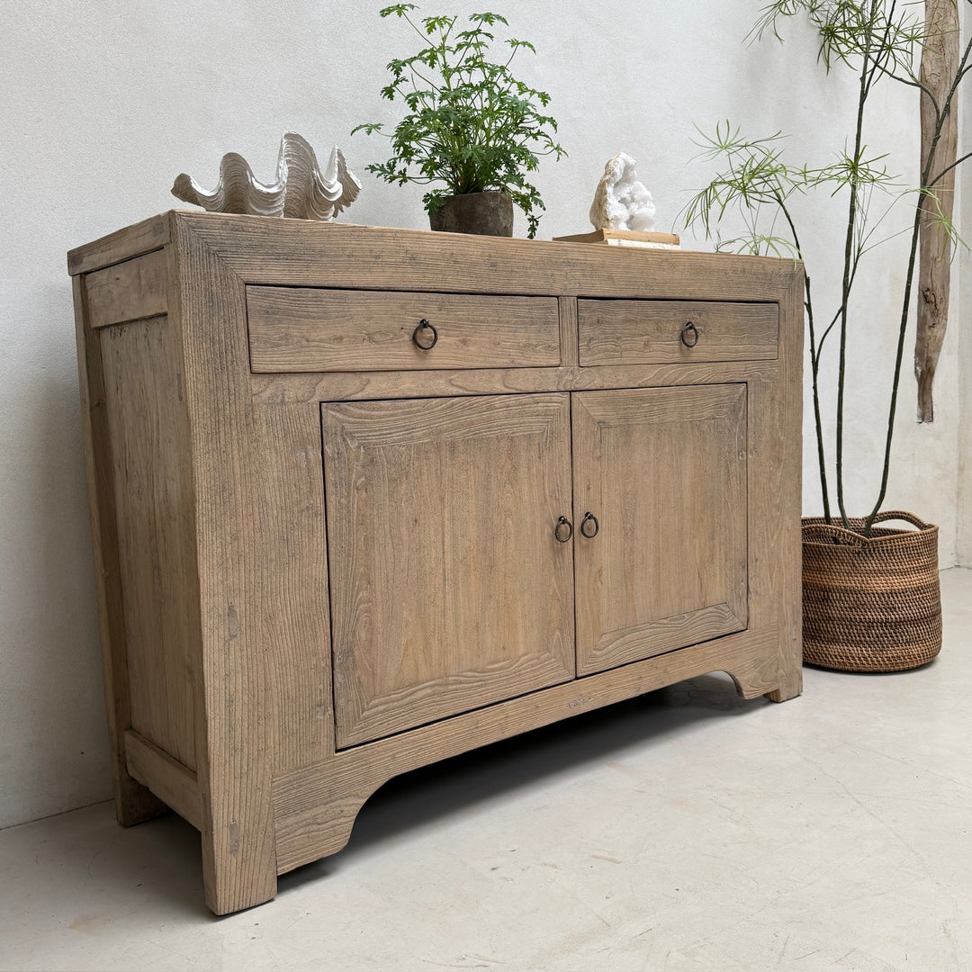 A rustic light brown wooden sideboard cabinet with two drawers on top and two paneled doors below, styled with small decorative figurines and potted plants in a neutral indoor setting.