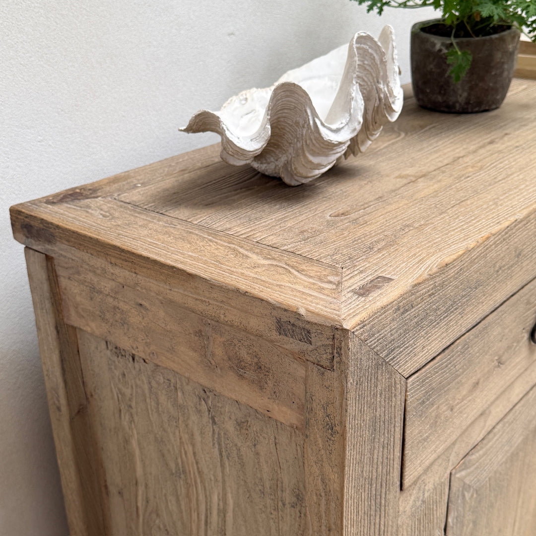 A rustic light brown wooden sideboard cabinet with a white clam shell sculpture and a potted plant on top, set against a plain off-white wall.