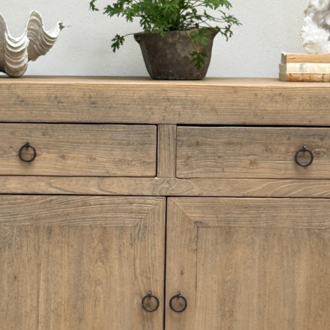 A rustic light brown wooden sideboard cabinet with two drawers on top and two doors below, each fitted with a small round black metal ring pull. The cabinet is styled in a home setting with a potted plant and decorative objects on top.