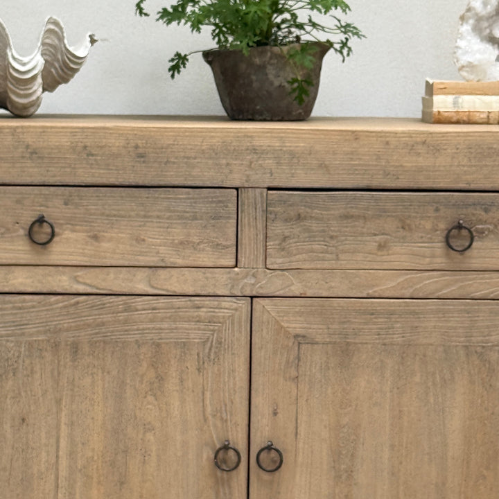 A rustic light brown wooden sideboard cabinet with two drawers on top and two doors below, each fitted with a small round black metal ring pull. The cabinet is styled in a home setting with a potted plant and decorative objects on top.
