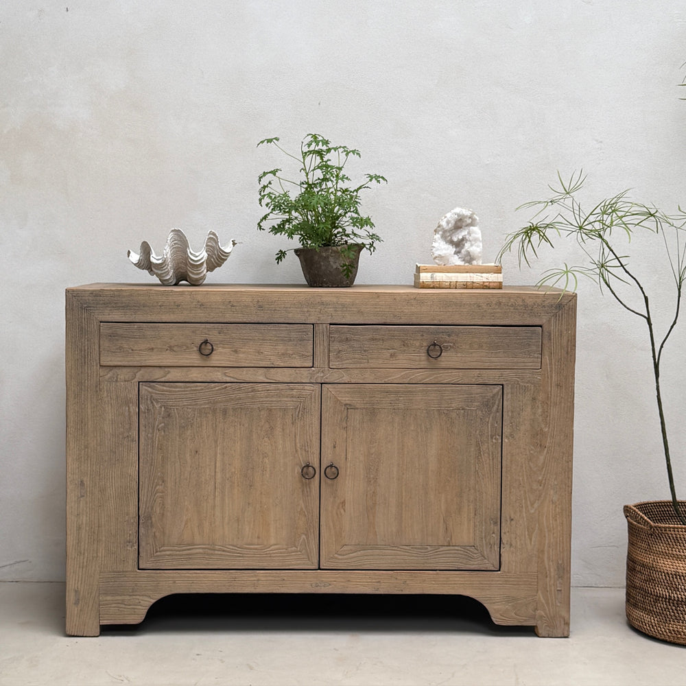 A rustic light brown wooden sideboard cabinet with two drawers and two doors, styled with decorative objects on top against a textured light gray wall.
