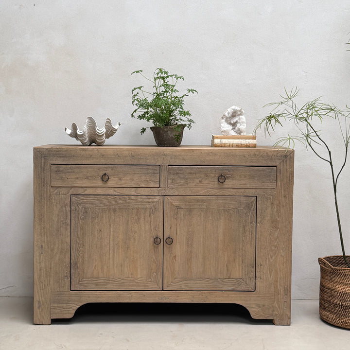 A rustic light brown wooden sideboard cabinet with two drawers and two doors, styled with decorative objects on top against a textured light gray wall.