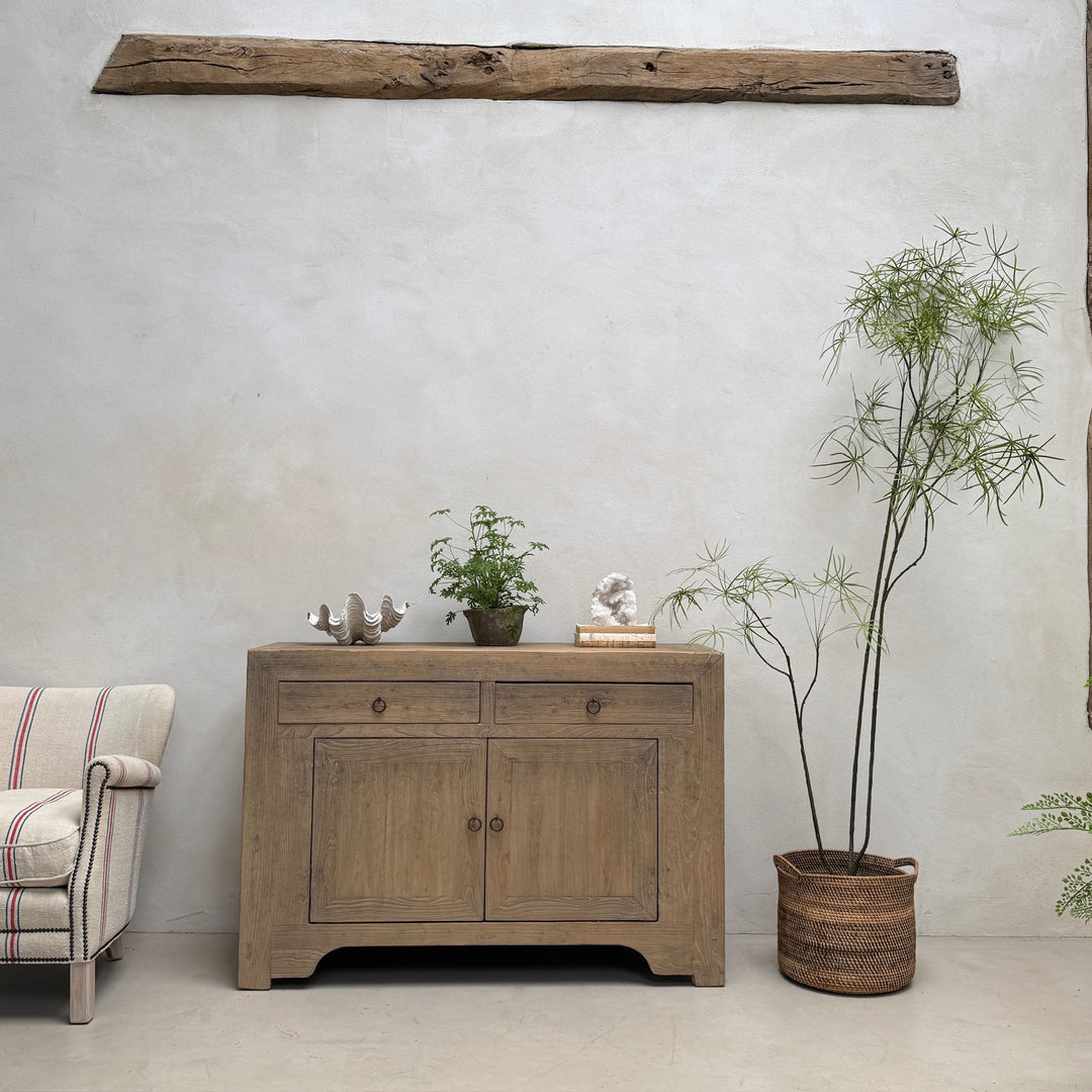 A rustic light brown wooden sideboard cabinet with two drawers and two doors, styled in a minimalist room setting with a textured white wall, a potted bamboo plant, and a striped armchair partially visible on the left.