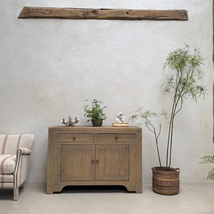 A rustic light brown wooden sideboard cabinet with two drawers and two doors, styled in a minimalist room setting with a textured white wall, a potted bamboo plant, and a striped armchair partially visible on the left.