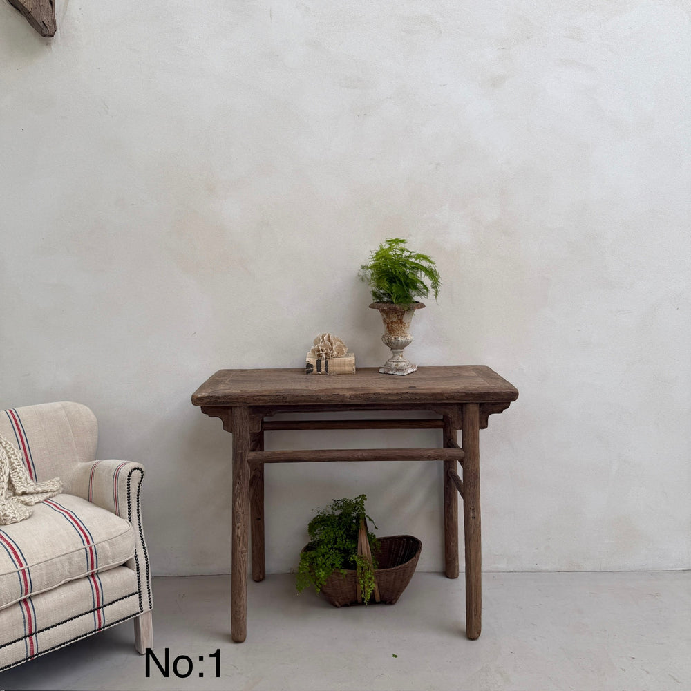 Wooden table with decorative items against a white wall, next to a plaid armchair.