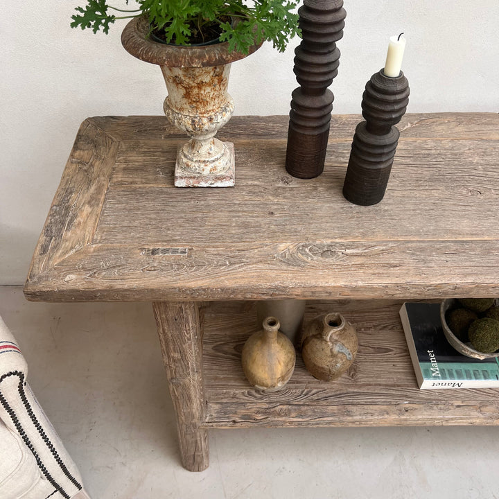 A rustic wooden coffee table with a distressed finish, styled with a potted green plant in a stone urn, dark wooden candle holders, and small decorative vases on the shelf.