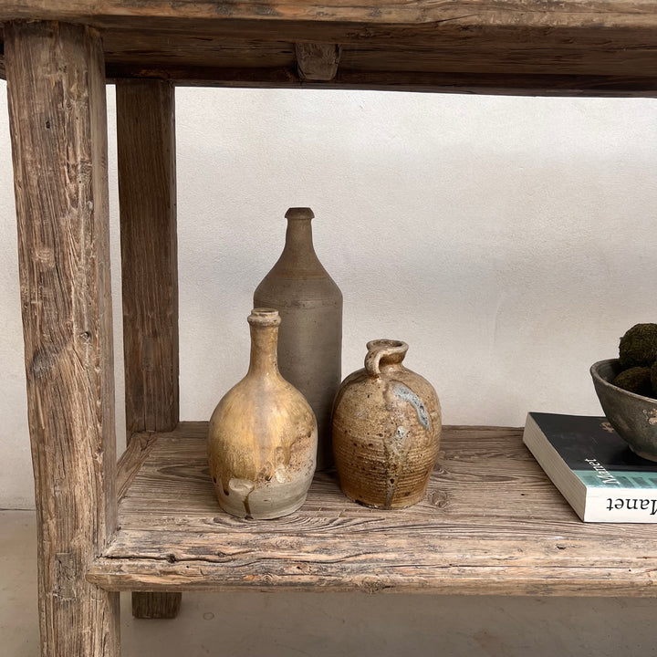 Three rustic handmade ceramic bottles and vases in earthy beige, brown, and gray tones displayed on a weathered wooden shelf.