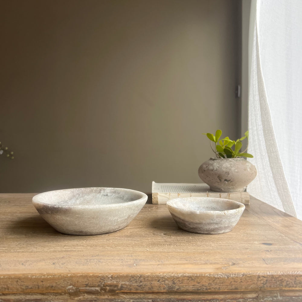 Two stone bowls on a wooden surface with a plant and books in the background.