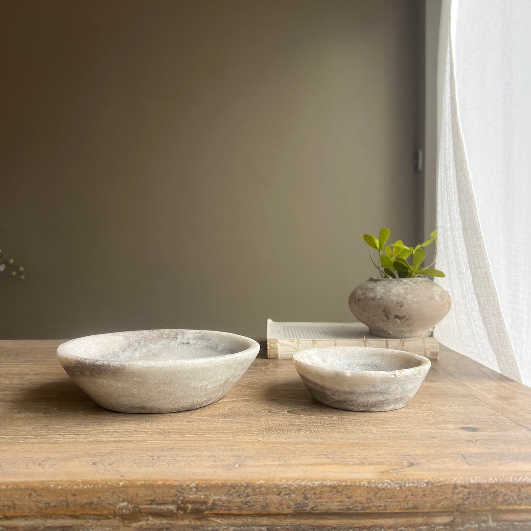 Two stone bowls on a wooden surface with a plant and books in the background.