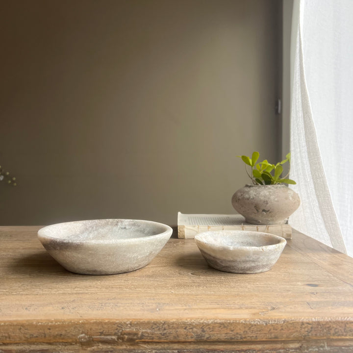 Two stone bowls on a wooden surface with a plant and books in the background.