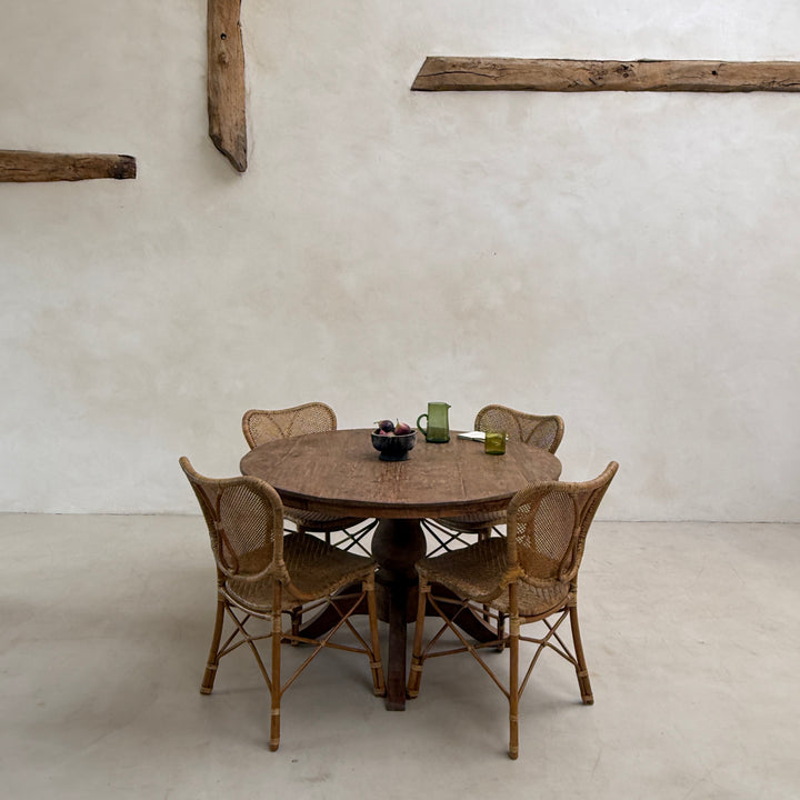 A round wooden dining table with four woven rattan chairs, set against a minimalist plaster wall with rustic wooden beams.