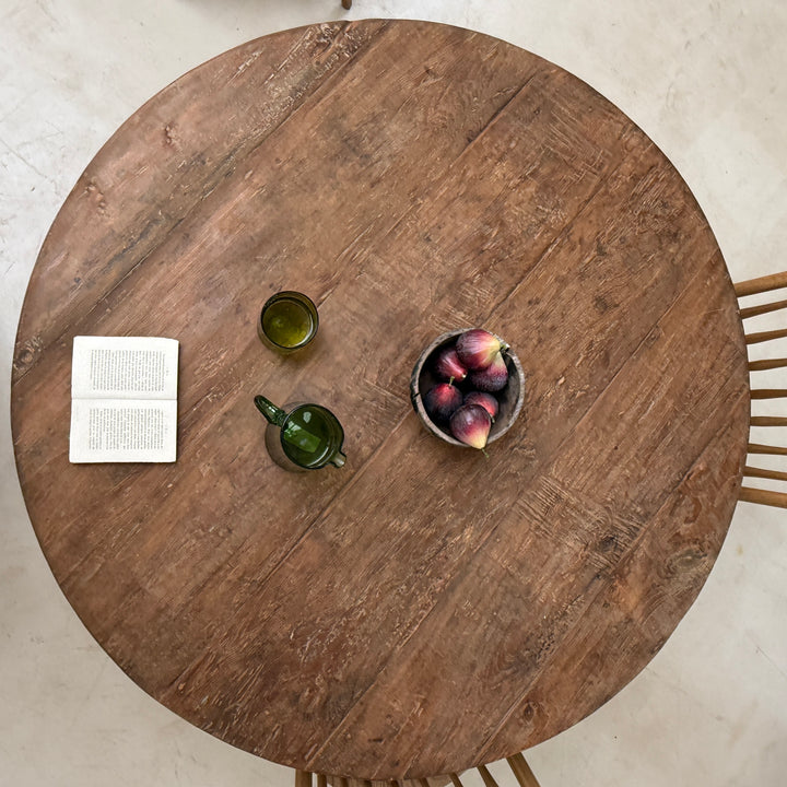 Overhead view of a round rustic wooden dining table with visible wood grain, styled with an open book, a small green glass cup, a green teapot, and a bowl of figs on a light stone floor.