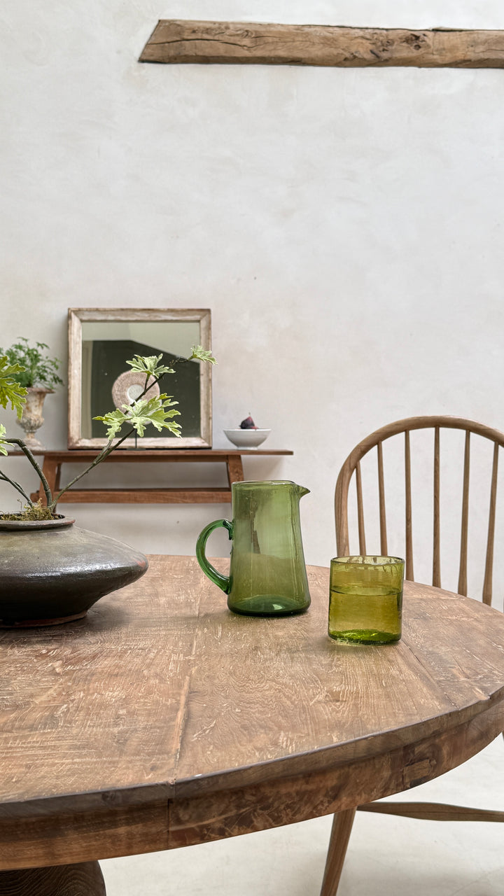 A green glass pitcher and matching tumbler set on a rustic wooden dining table, with a wooden chair and a neutral-toned wall in the background.