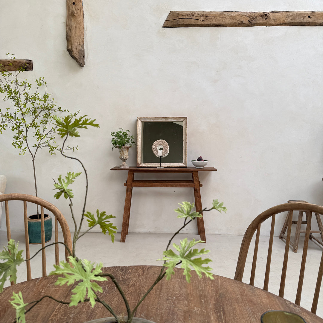 A styled interior scene with a round wooden dining table in the foreground, wooden chairs, potted plants, and a small wooden console table against a textured off-white wall. A framed circular artwork sits on the console table.