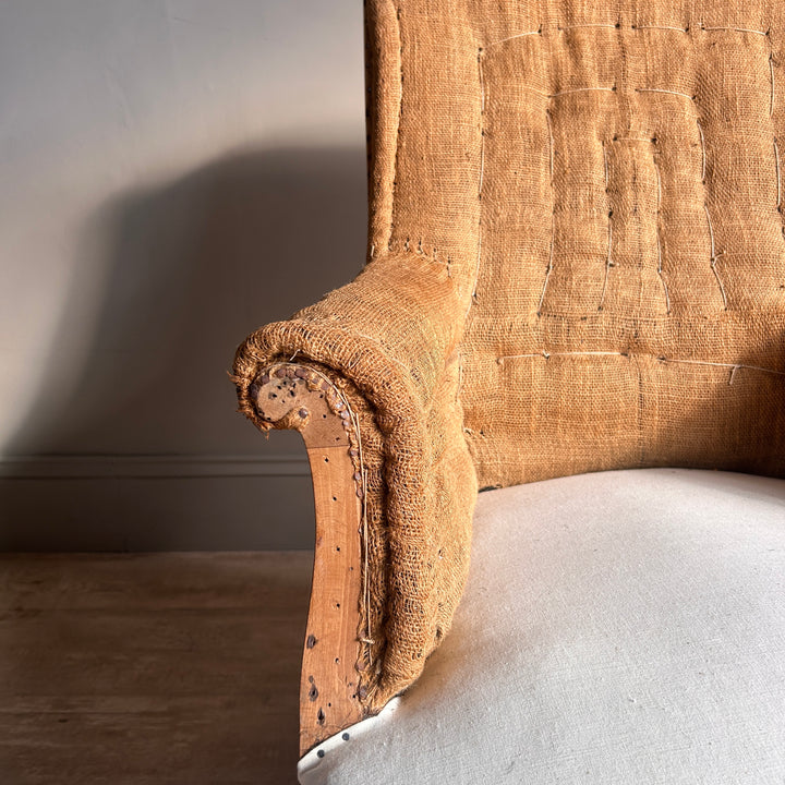 Close-up of the beautifully shaped rolled arm of an antique French chair, revealing aged timber, original upholstery tacks and textured hessian fabric.