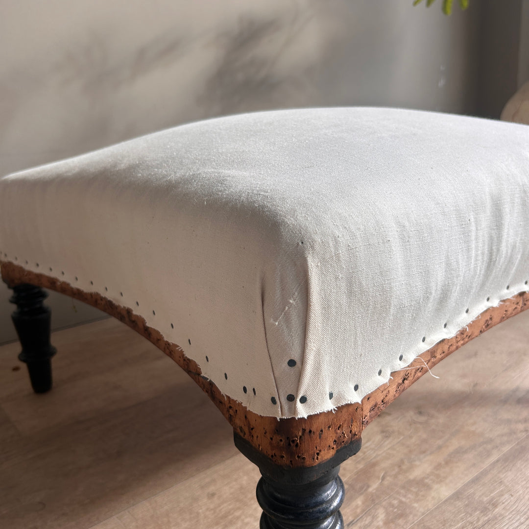 Close-up of an antique French footstool showing its cream calico upholstery, gently curved timber frame, visible upholstery tacks and turned wooden leg.