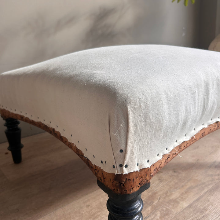 Close-up of an antique French footstool showing its cream calico upholstery, gently curved timber frame, visible upholstery tacks and turned wooden leg.