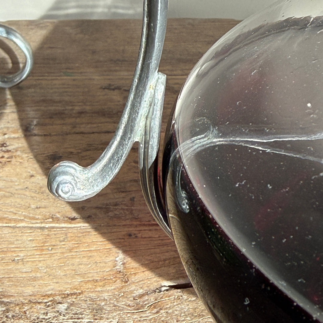 Close-up of the elegant curved pewter handle and decorative scroll detailing beside the rounded glass body of a French wine decanter.