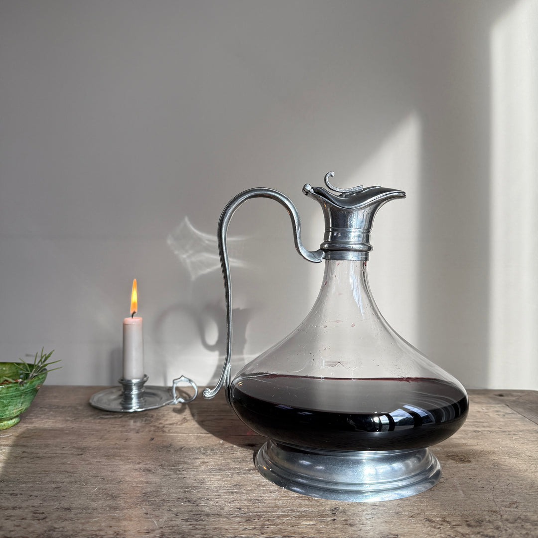 Traditional French pewter and glass red wine decanter with an ornate handle and rounded body, styled on a rustic table beside a lit candle.