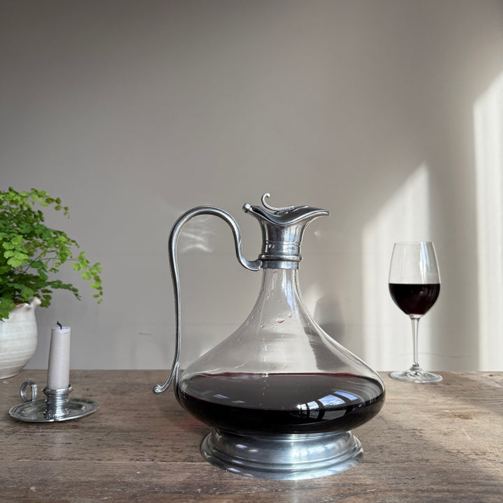 French pewter and glass wine carafe displayed on a rustic wooden dining table alongside a glass of red wine and a candle.