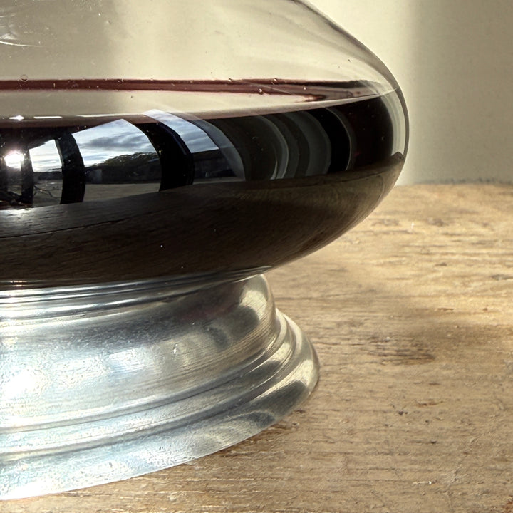 Close-up of the rounded clear glass body of a French wine decanter, showing red wine resting above its decorative pewter pedestal base.