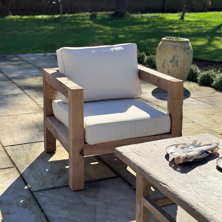 Wooden outdoor chair with white cushions on a patio, surrounded by greenery.